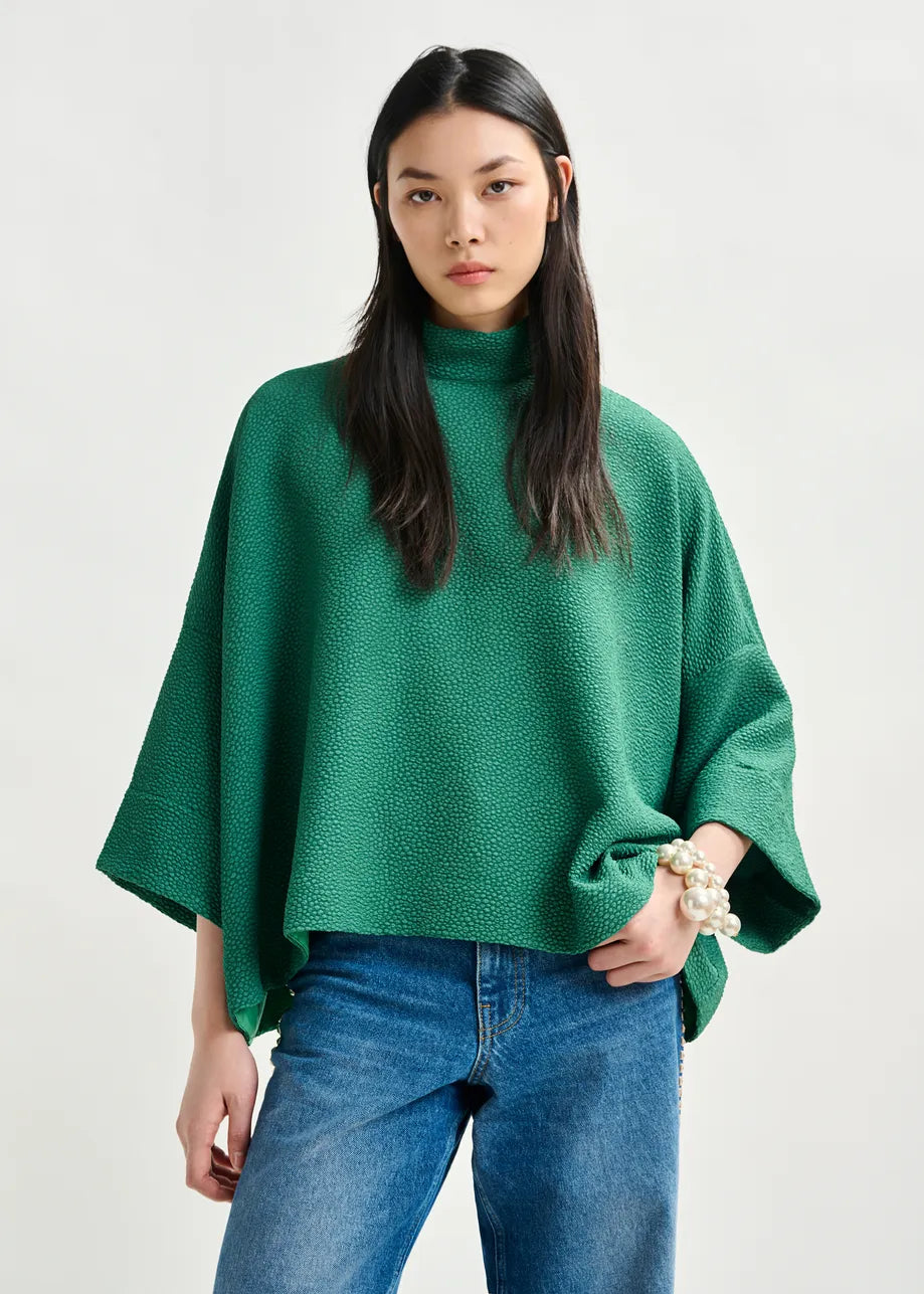 A woman models the Iscratch Oversized Top in green by Essentiel Antwerp, styled with blue jeans and a chunky pearl bracelet, standing against a plain light background.
