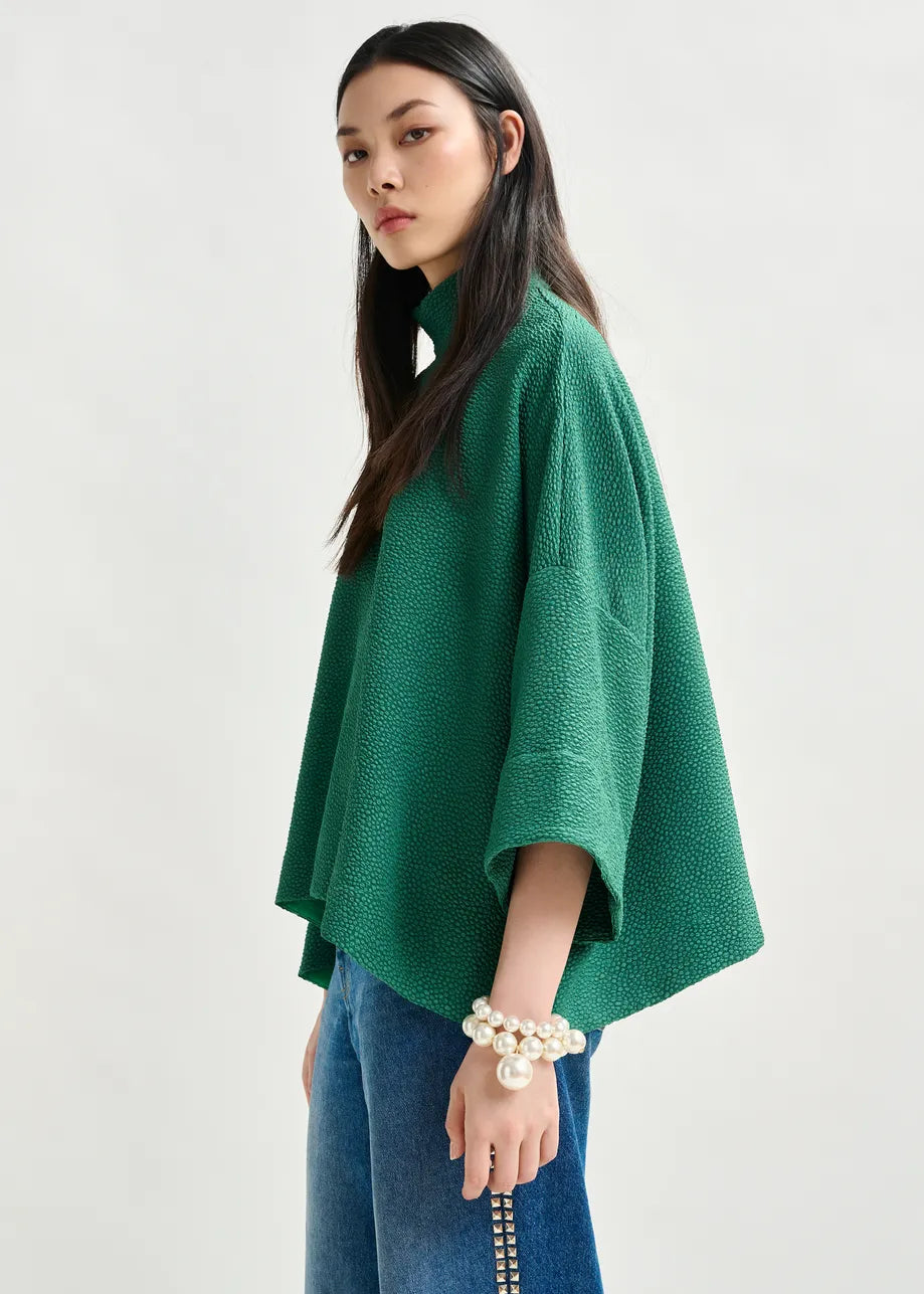A woman stands sideways against a plain light background, wearing the Essentiel Antwerp Iscratch Oversized Top in green and wide blue jeans, styled with pearl bracelets.