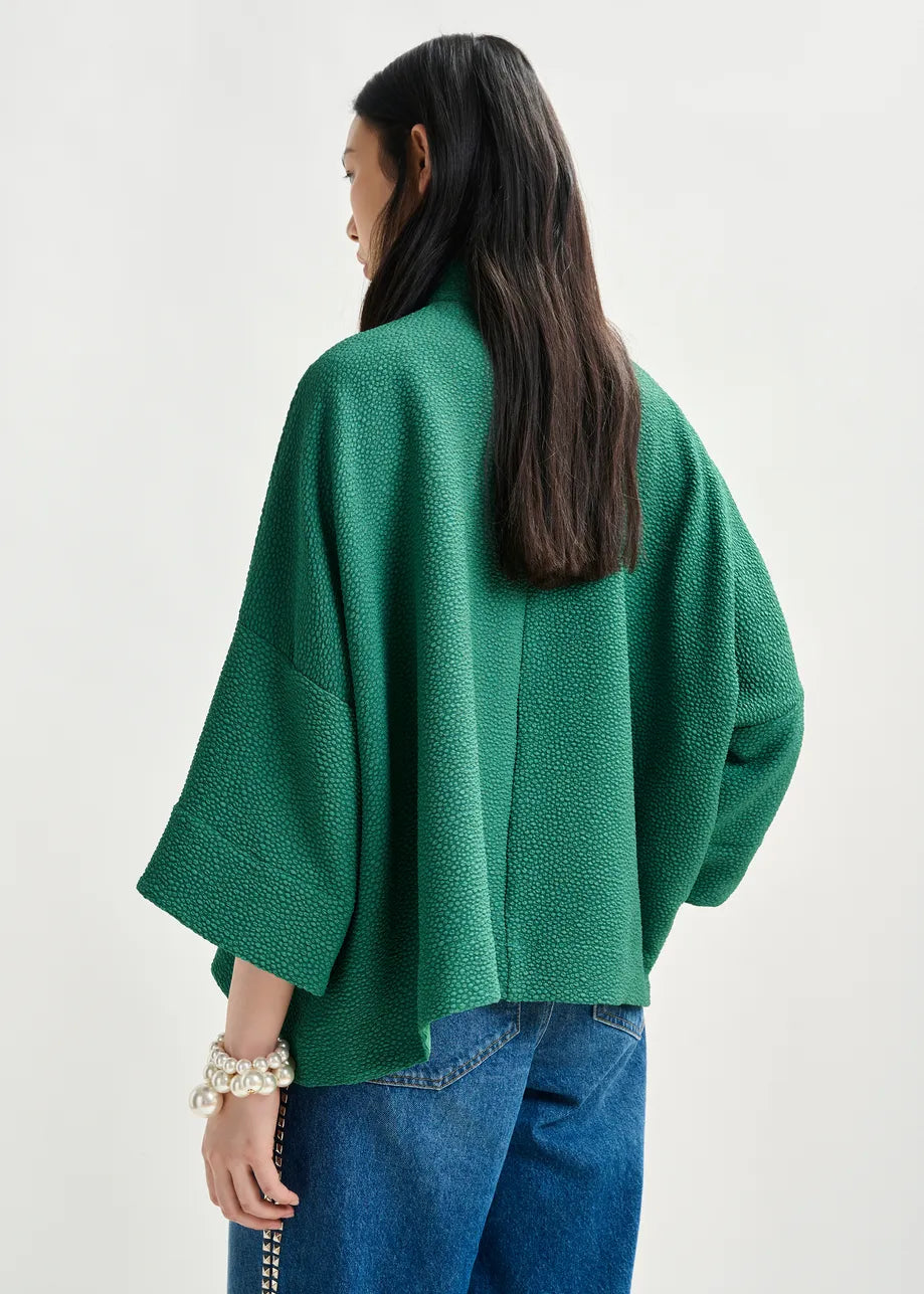 A woman with long dark hair, wearing the Essentiel Antwerp Iscratch Oversized Top in green and blue jeans, stands with her back to the camera against a plain light background.
