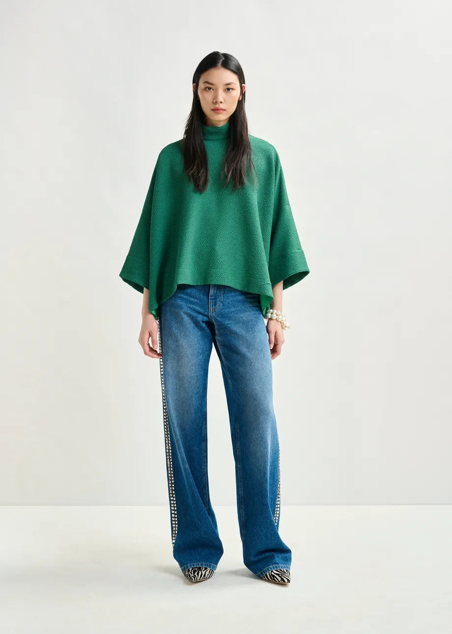 A woman faces forward in the Essentiel Antwerp Iscratch Oversized Top in green, paired with wide-leg embellished jeans and pointed shoes, set against a plain light background.