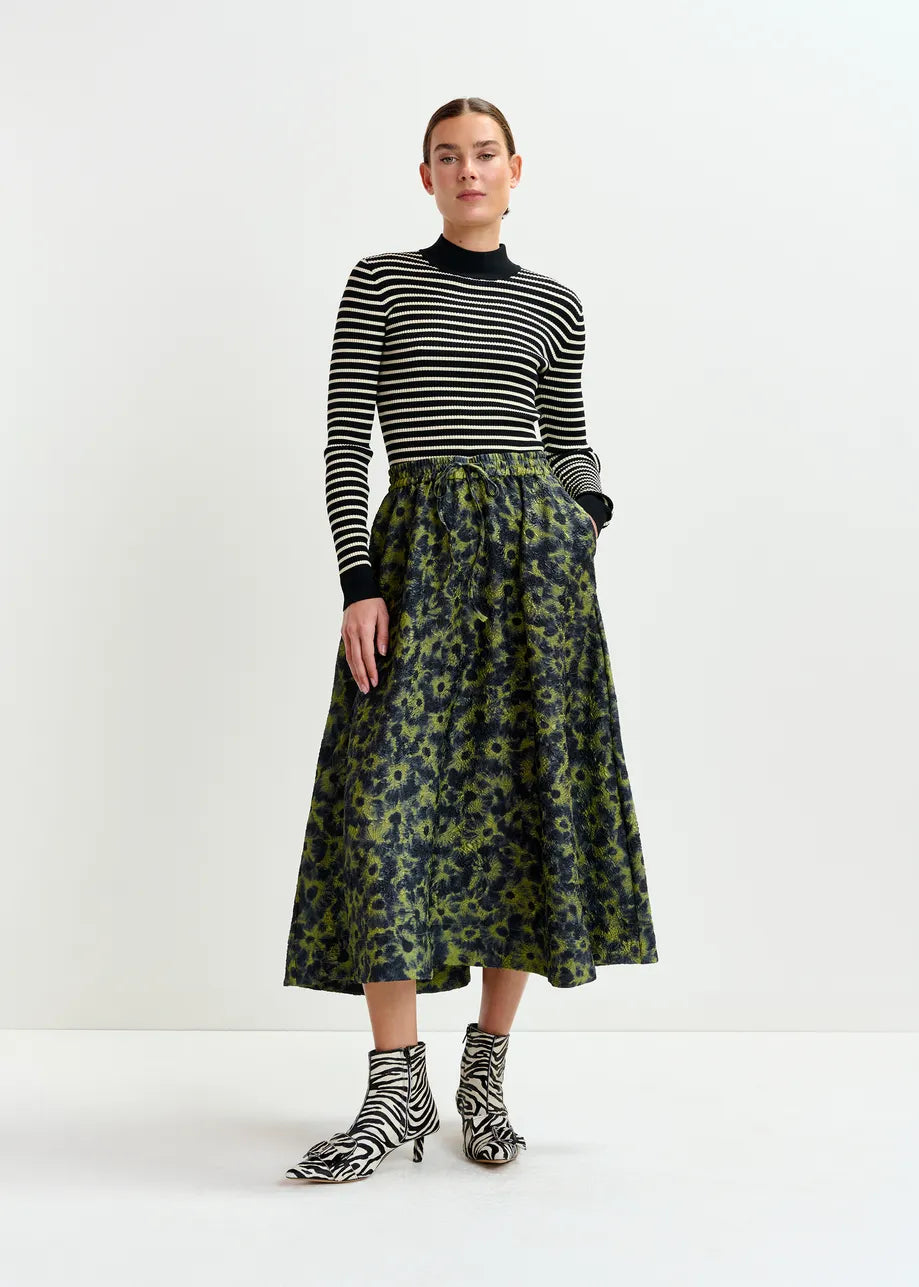 A person stands against a white background, wearing a black and white striped top, Essentiel Antwerp’s Isebel Floral Jacquard Skirt in khaki, and matching zebra-print ankle boots.