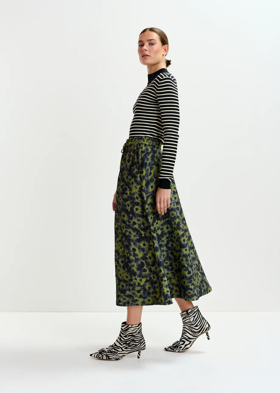 Woman in profile wears a black-and-white striped turtleneck, Essentiel Antwerp's Isebel Floral Jacquard Skirt in khaki, and black-and-white zebra print heeled boots.