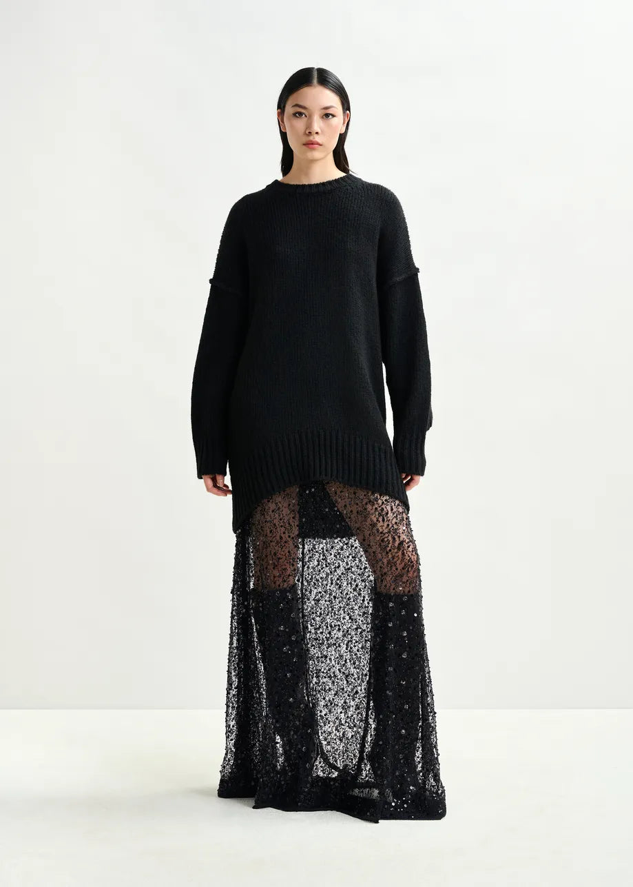 A woman stands against a plain background wearing an Essentiel Antwerp Isequin Embellished Maxi Skirt in black, paired with a long black sweater.