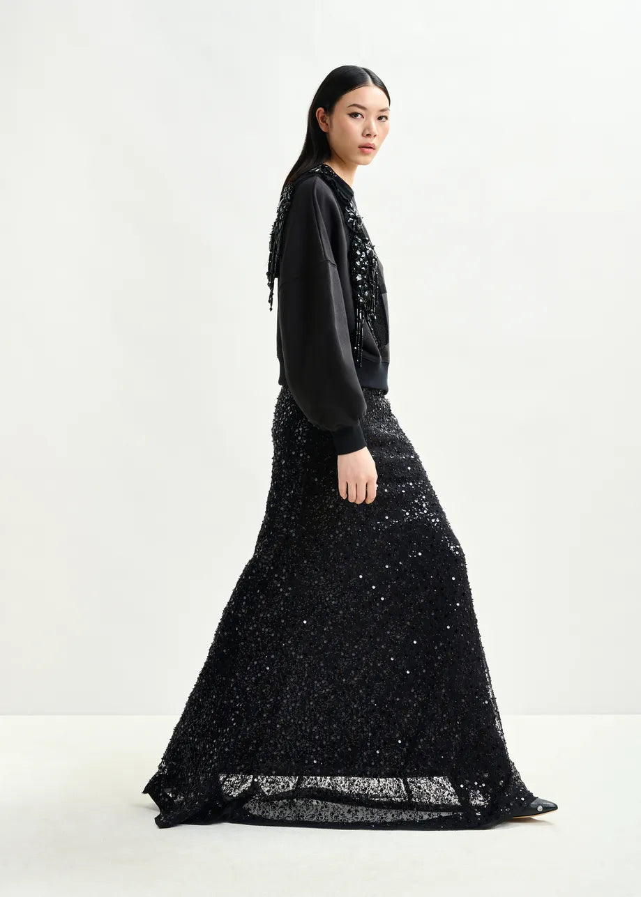 A woman stands sideways against a light background, wearing the Essentiel Antwerp Isequin Embellished Maxi Skirt in black paired with a matching sequin-embellished black sweatshirt.