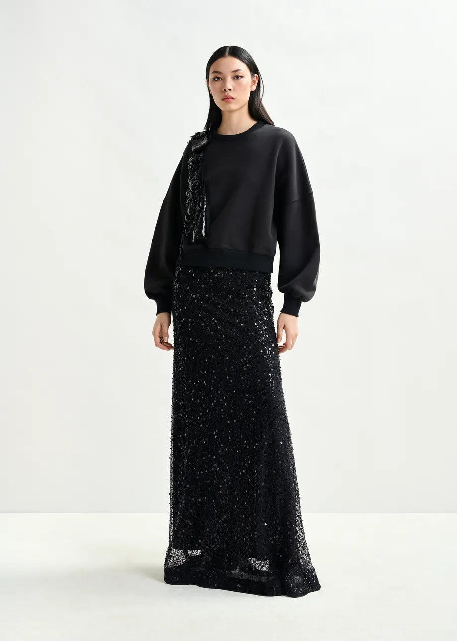 A woman wears the Essentiel Antwerp Isequin Embellished Maxi Skirt in black with a black balloon-sleeve sweatshirt, posed against a plain light background.