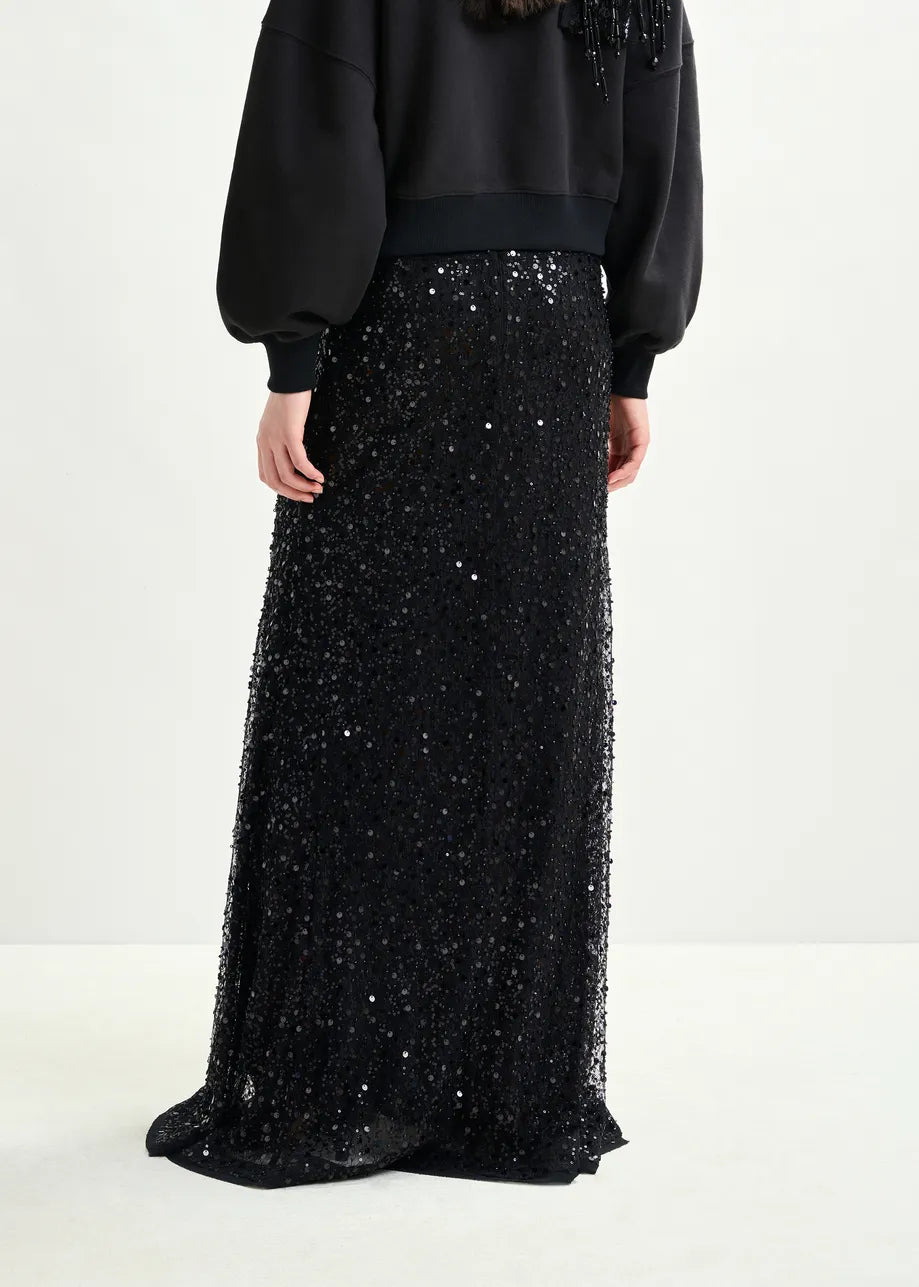 A person wearing the Essentiel Antwerp Isequin Embellished Maxi Skirt in black and a black sweatshirt, photographed from the back against a plain light background.