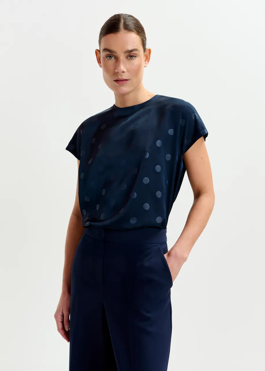 A woman models the Essentiel Antwerp Ishirt Polkadot Top in navy, paired with matching pants, standing against a plain light background with one hand in her pocket.