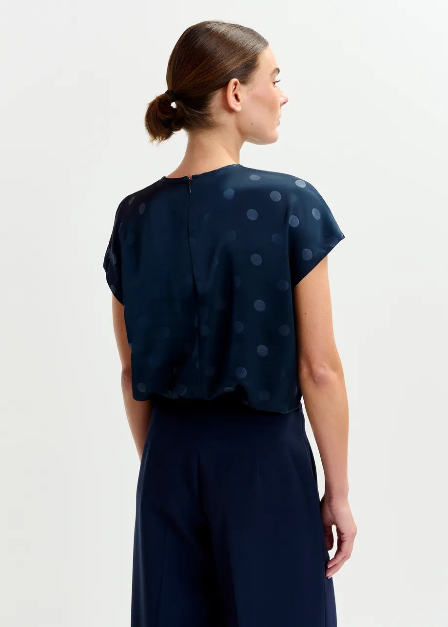A woman faces away, wearing the Essentiel Antwerp Ishirt Polkadot Top in navy with matching dark trousers, her hair tied back in a low bun.
