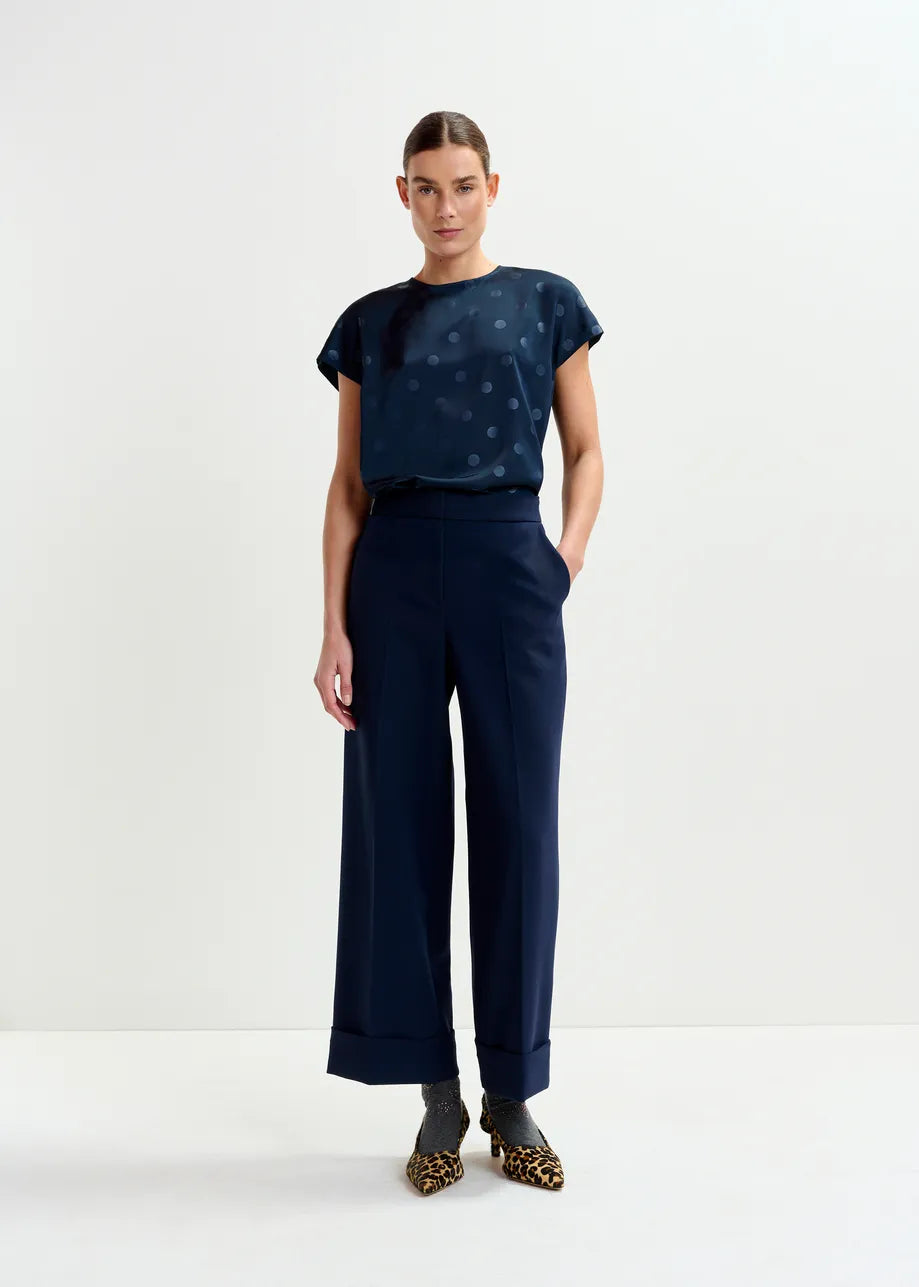 A woman stands against a plain background wearing the Essentiel Antwerp Ishirt Polkadot Top in navy, wide-leg navy trousers, gray socks, and leopard-print shoes.