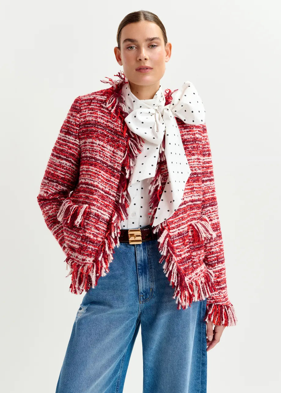 A woman in the Essentiel Antwerp Iskra Bouclé Jacket in burgundy red, styled with a white blouse with a polka dot bow and blue jeans, stands against a plain background.