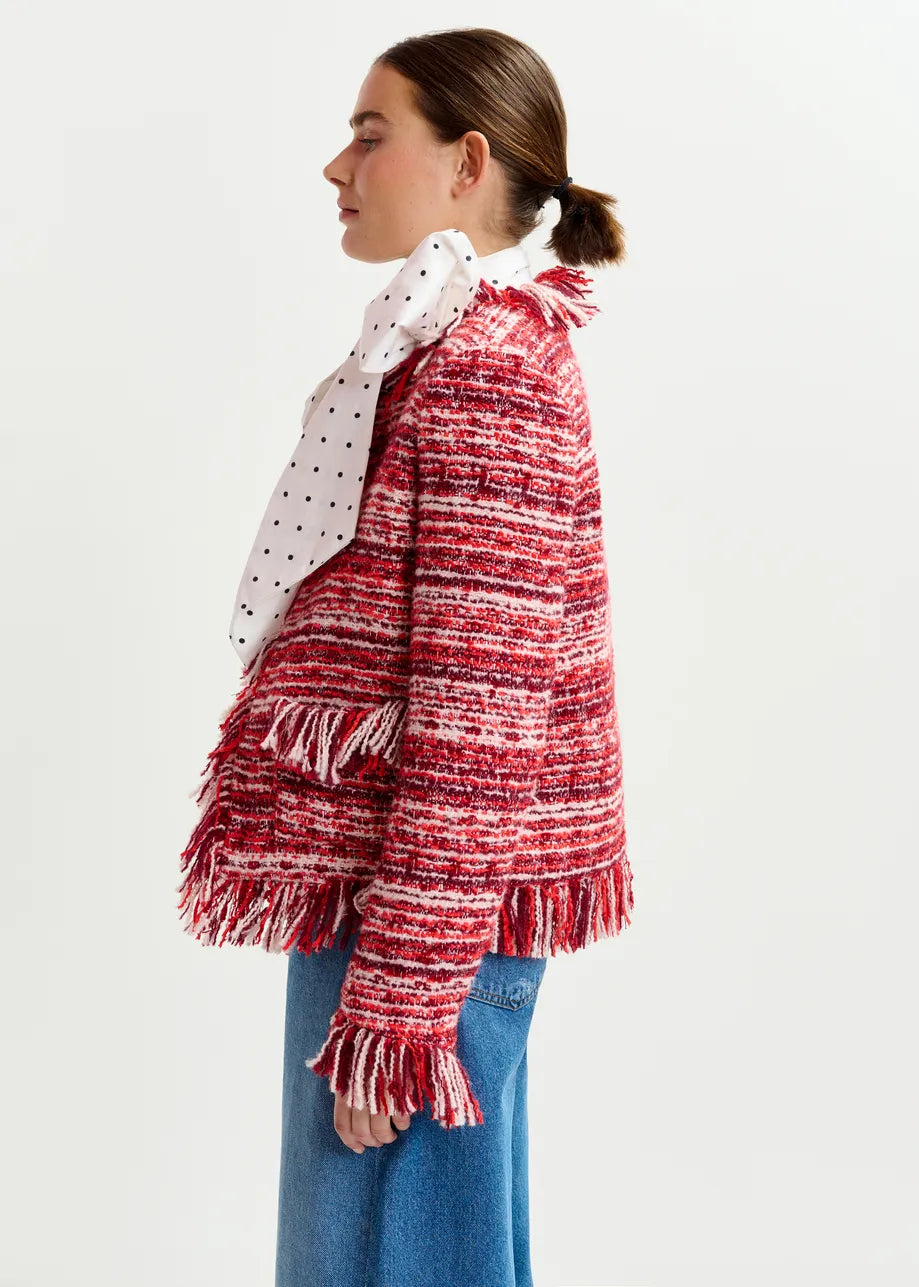 A woman in profile wears the Essentiel Antwerp Iskra Bouclé Jacket in burgundy red, styled with blue jeans and a white scarf with black polka dots, standing against a plain background.