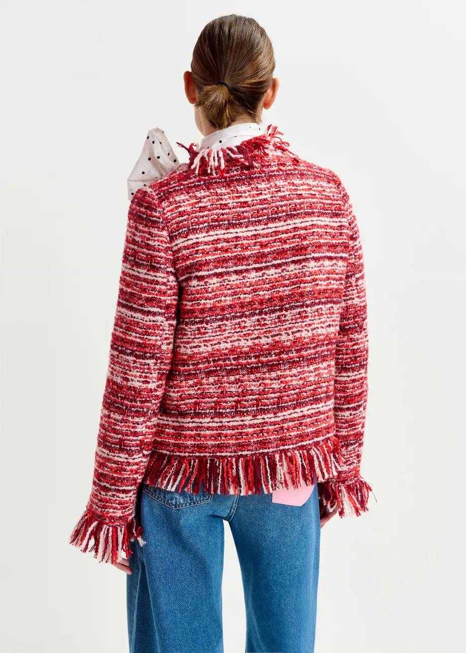 A person stands facing away, wearing the Essentiel Antwerp Iskra Bouclé Jacket in Burgundy Red, featuring a boxy silhouette and red-and-white fringe, styled with blue jeans and a white polka-dot collar shirt.