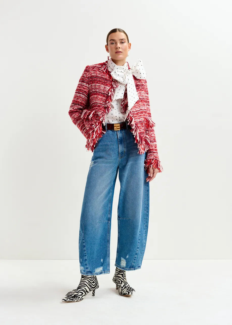 A person stands against a plain background wearing the Iskra Bouclé Jacket in Burgundy Red by Essentiel Antwerp, a polka dot blouse with a bow, wide-leg blue jeans, and zebra print boots.