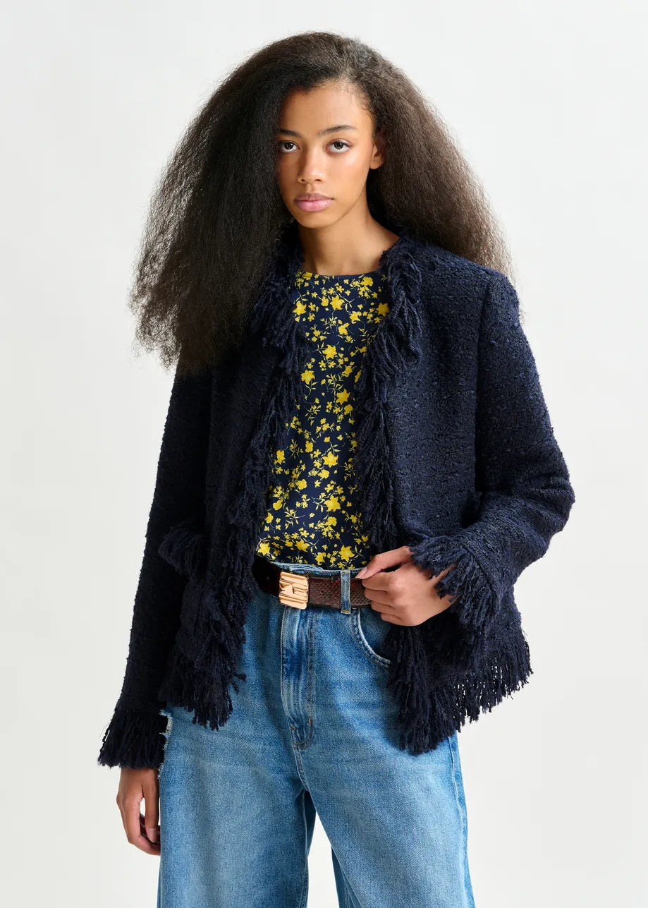A woman with long, voluminous hair wears the Essentiel Antwerp Iskra Bouclé Jacket in navy over a black and yellow floral top, blue jeans, and a brown belt, standing against a plain light background.