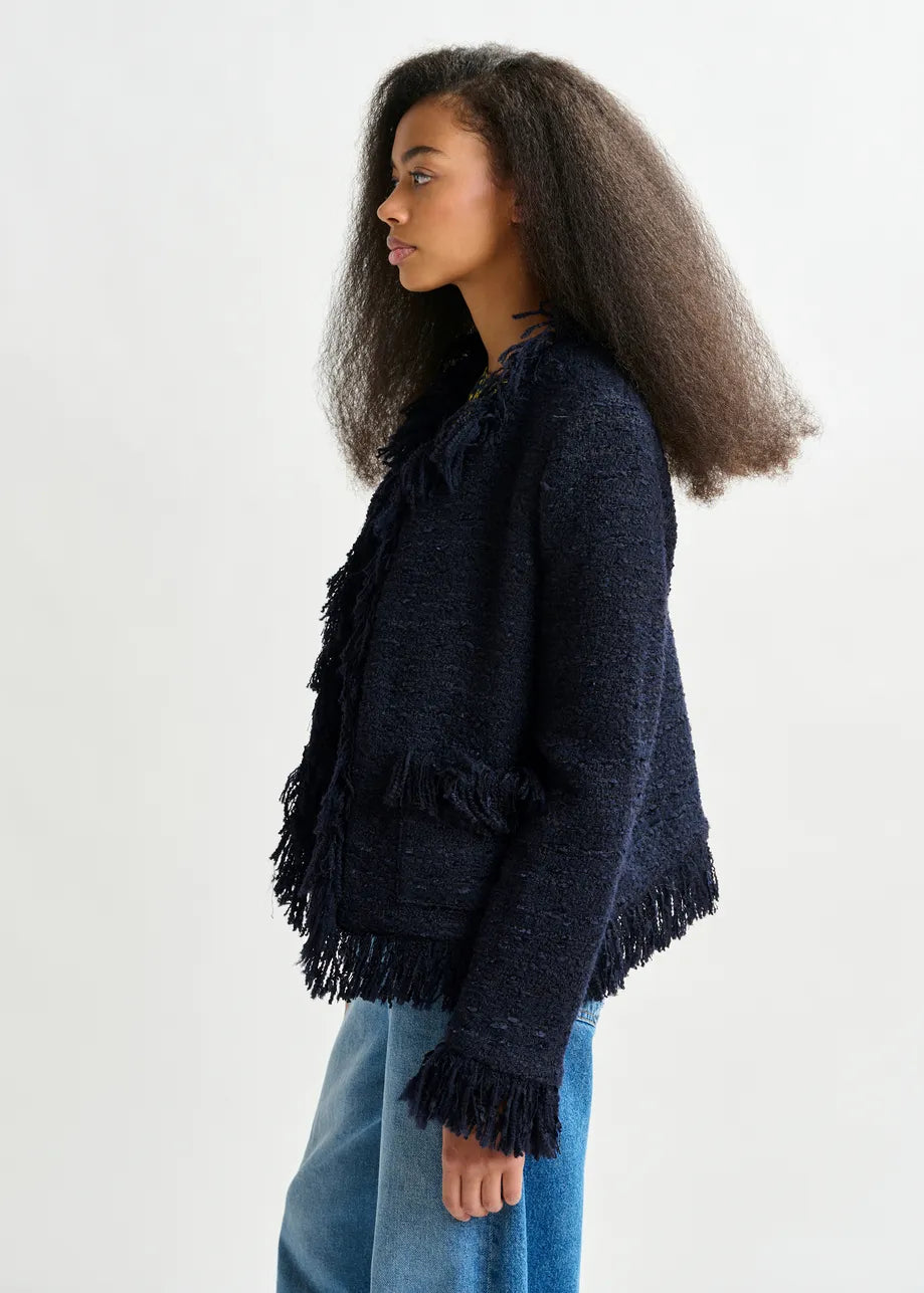 A woman with long, voluminous hair poses in profile against a plain light background, wearing Essentiel Antwerp's Iskra Bouclé Jacket in navy with fringe detailing, paired with blue jeans.