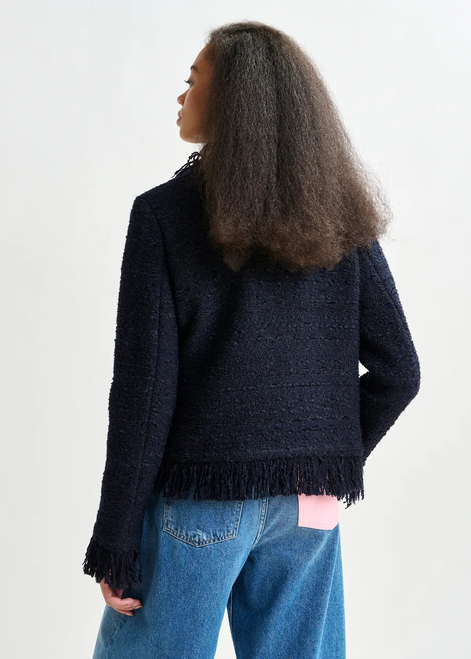 A person with long, textured hair wears the Essentiel Antwerp Iskra Bouclé Jacket in navy, featuring fringe at the hem and cuffs, styled with blue jeans and a pink shirt peeking out at the back.
