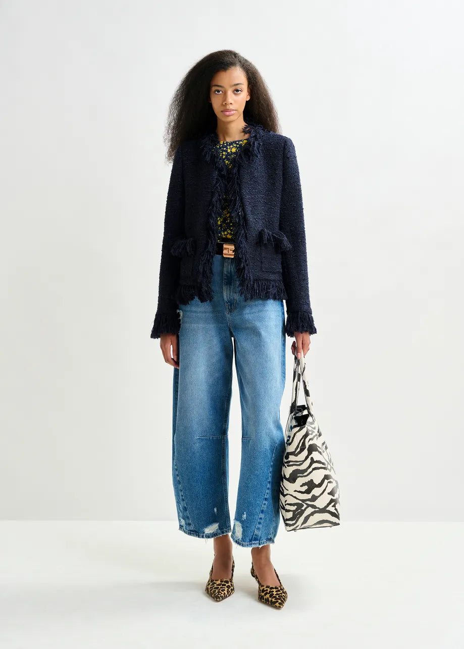 A woman stands against a plain background wearing the Essentiel Antwerp Iskra Bouclé Jacket in navy, styled with wide-leg blue jeans, animal print heels, and a zebra print tote bag.