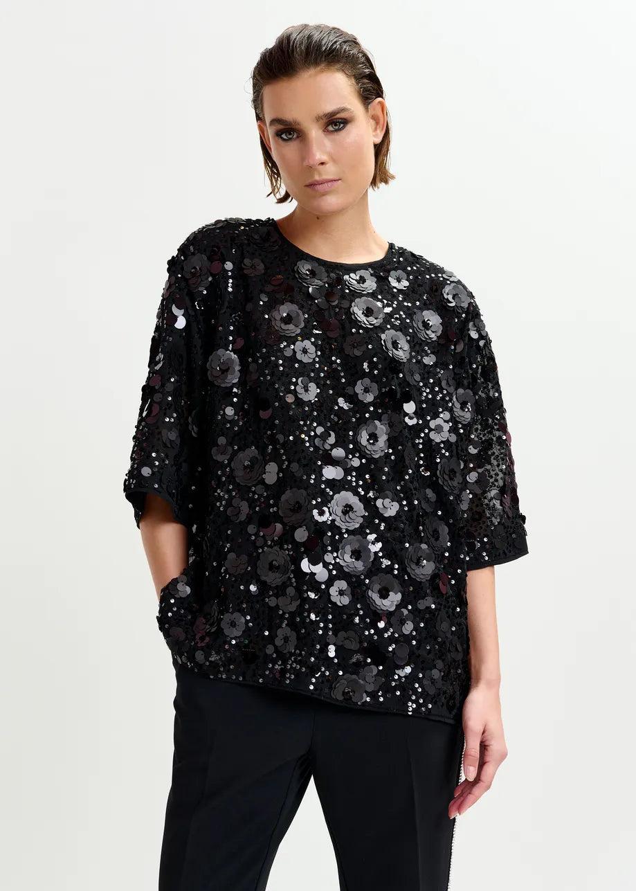 A person wears the Essentiel Antwerp Isparkle Sequin-Embellished Top in black with black pants, standing against a plain light background.