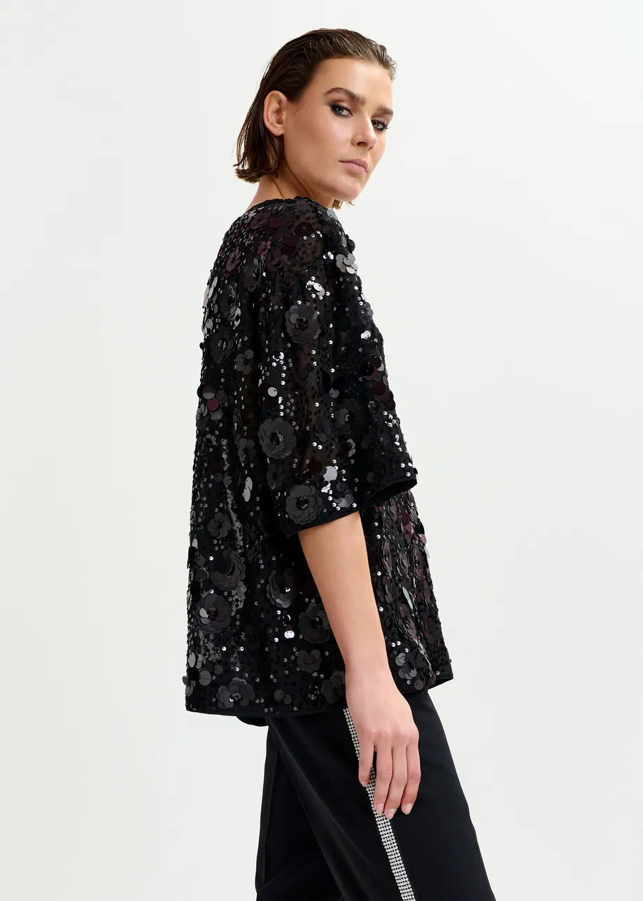 A person stands sideways in a loose, boxy Essentiel Antwerp Isparkle Sequin-Embellished Top in black, paired with striped black pants, against a plain light background.