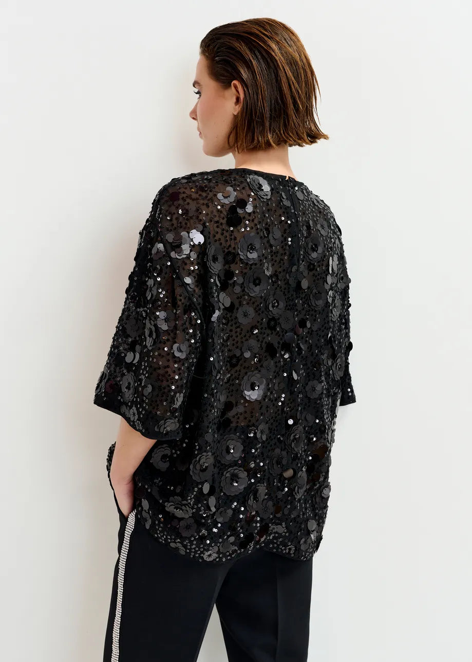 A person stands facing away against a white background, wearing the Essentiel Antwerp Isparkle Sequin-Embellished Top in black with a boxy silhouette and short sleeves, paired with black pants featuring a side stripe.