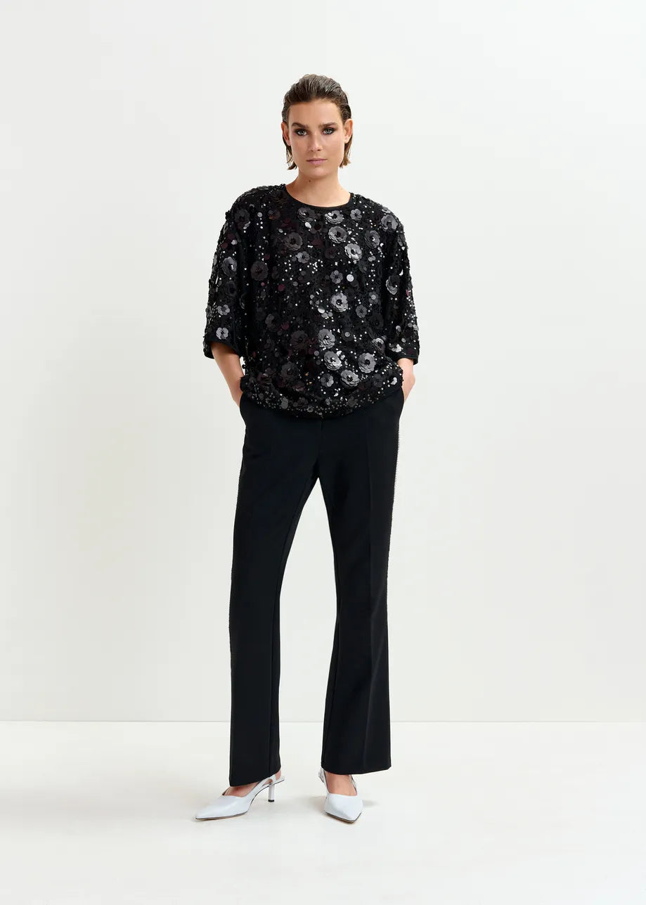 A person stands against a plain background wearing an Essentiel Antwerp Isparkle Sequin-Embellished Top in black, paired with black trousers and white heels.