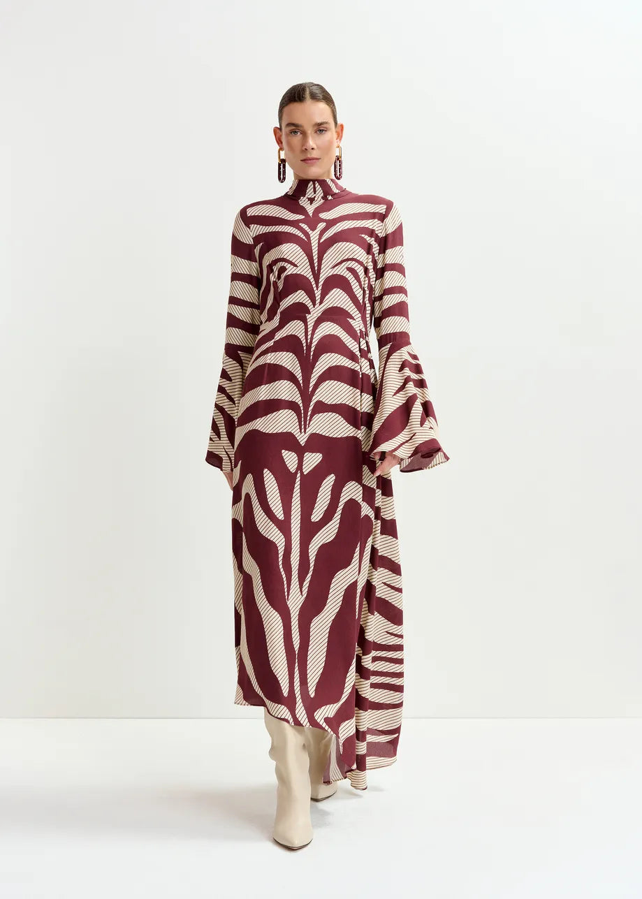 A woman stands against a white background wearing the Essentiel Antwerp Istripe Dress in burgundy, featuring a high neck and flared sleeves, paired with white knee-high boots.