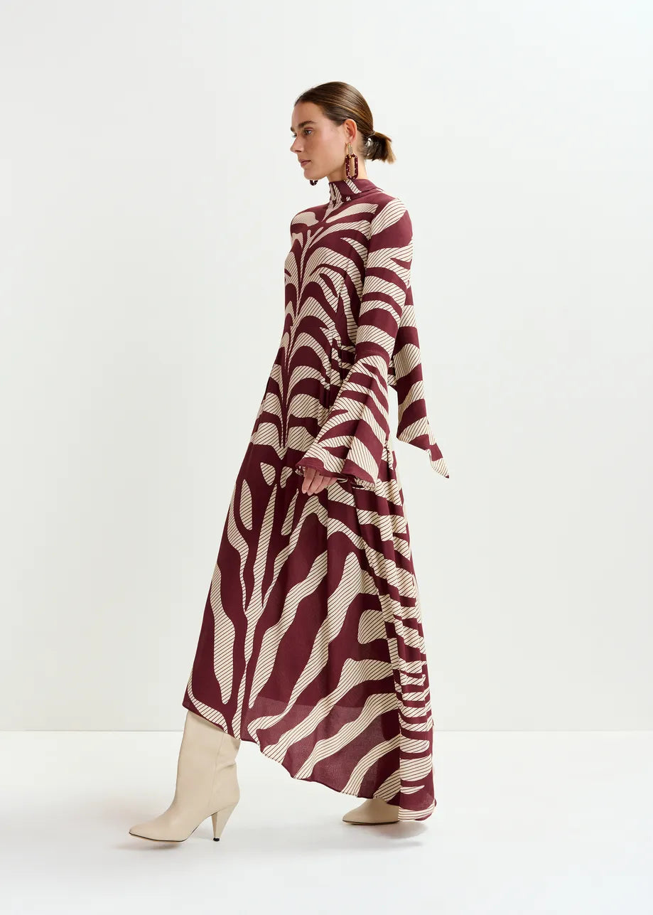 A woman stands in profile against a white background, wearing the Essentiel Antwerp Istripe Dress in burgundy with flared sleeves, cream heeled boots, and statement earrings.