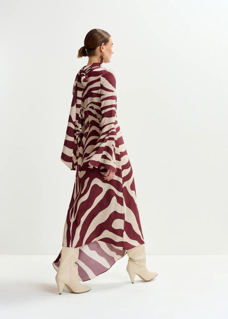 A woman in the Essentiel Antwerp Istripe Dress—burgundy with cream patterns, flared sleeves—pairs it with cream knee-high boots, posing against a plain white background.