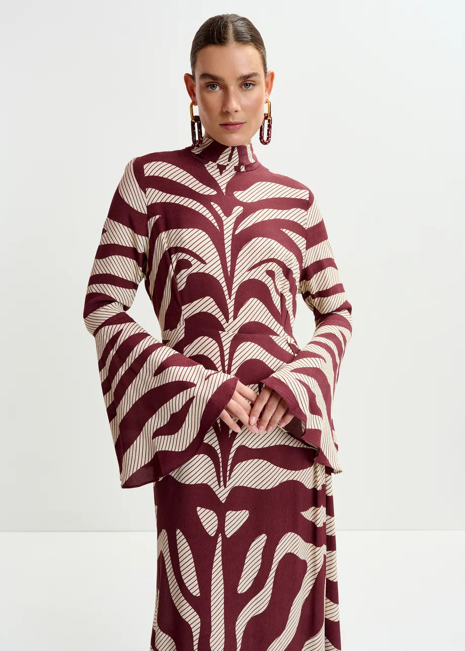A woman models the Essentiel Antwerp Istripe Dress in burgundy, featuring a high neck and abstract cream patterns, against a plain white background.
