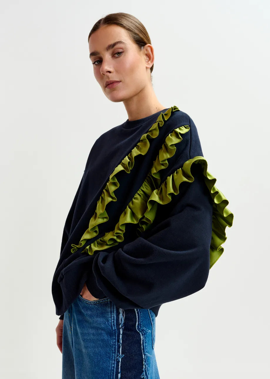 A person wearing the Essentiel Antwerp Iwave Ruffle Sweatshirt in navy, featuring green sleeve details, and blue jeans stands against a plain white background.