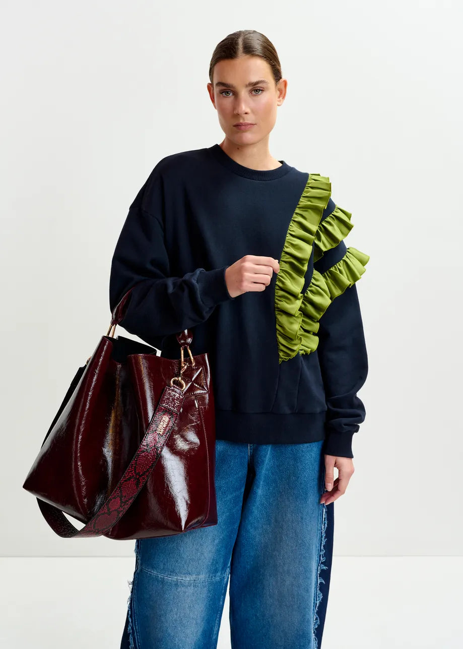 A woman wearing the Essentiel Antwerp Iwave Ruffle Sweatshirt in navy, paired with loose blue jeans and carrying a large burgundy handbag with a patterned strap, stands against a plain white background.