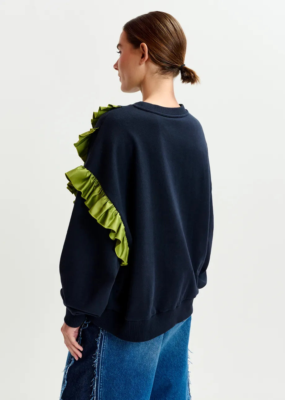 A person wears the Essentiel Antwerp Iwave Ruffle Sweatshirt in navy, featuring asymmetric ruffles with green accents on the shoulder and sleeve, paired with blue jeans and standing against a plain background.