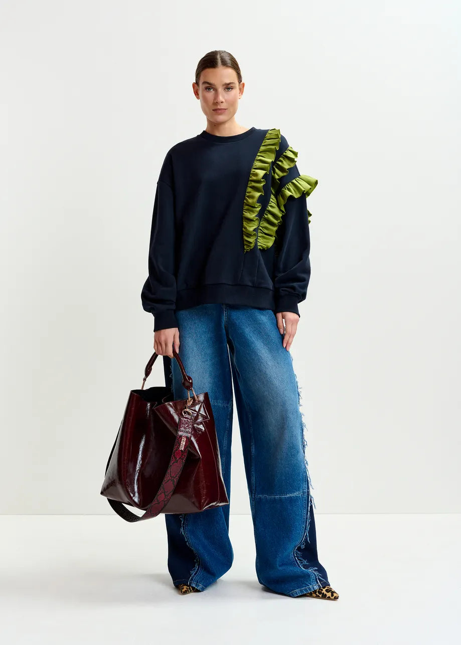 A woman stands against a white background wearing the Essentiel Antwerp Iwave Ruffle Sweatshirt in navy, styled with wide-leg blue jeans, leopard print shoes, and carrying a large burgundy handbag.