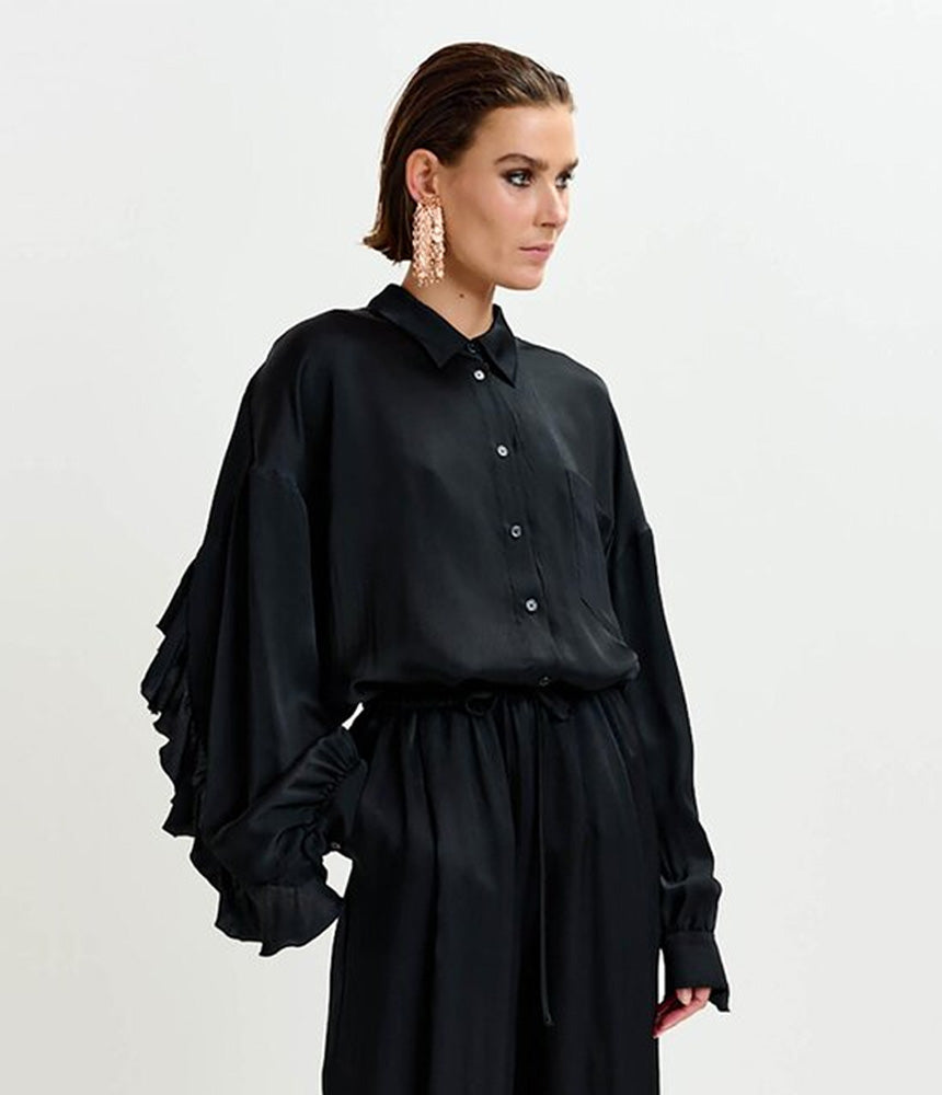 A woman stands against a plain white background wearing Essentiel Antwerp's Irving Wide Trousers in black and a loose-fitting black button-up blouse with ruffled sleeves.