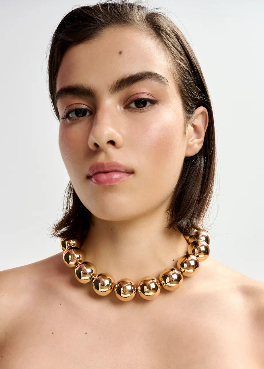 A person with short brown hair wears the Jadine Gold Tone Necklace by Essentiel Antwerp, a chunky statement piece featuring metallic-tone beads, against a plain light background.