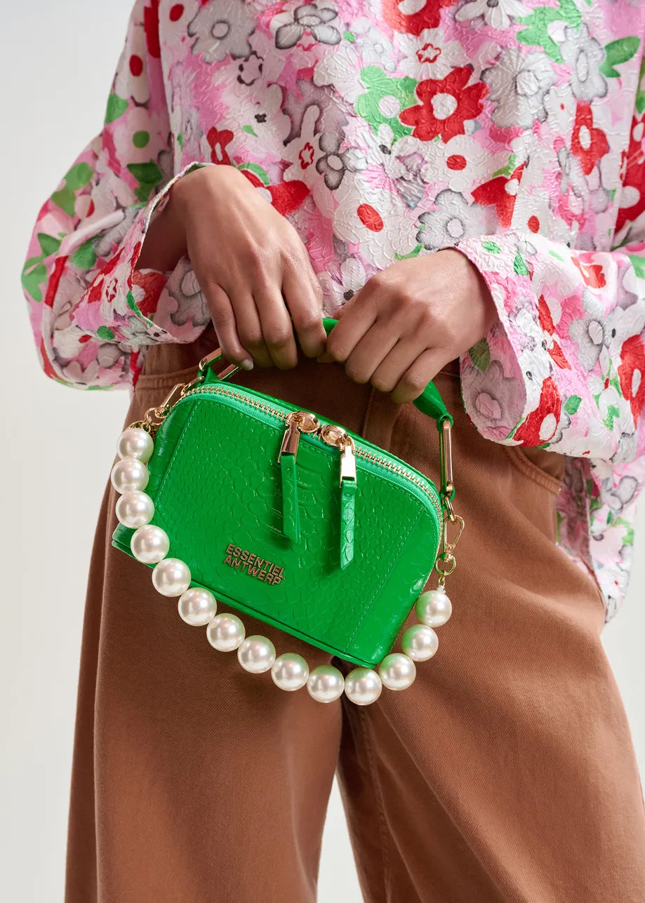 A person wearing a pink floral blouse and brown pants holds a small green handbag with gold zippers and a large pearl handle, styled with the Essentiel Antwerp Jadine Gold Tone Necklace – Pearl for a chic, versatile look.