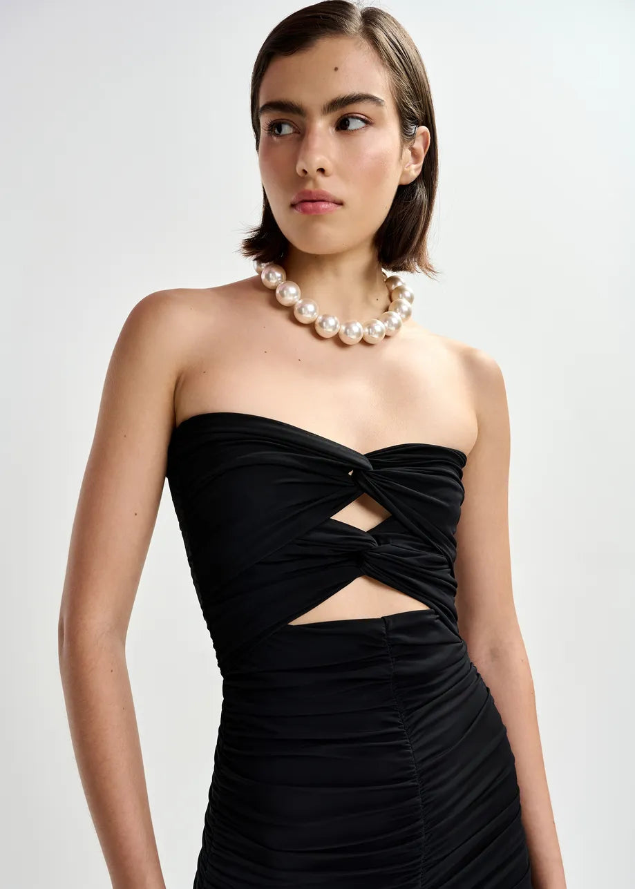 A woman in a strapless black dress with cutouts wears the Essentiel Antwerp Jadine Gold Tone Necklace - Pearl, standing against a plain light background.