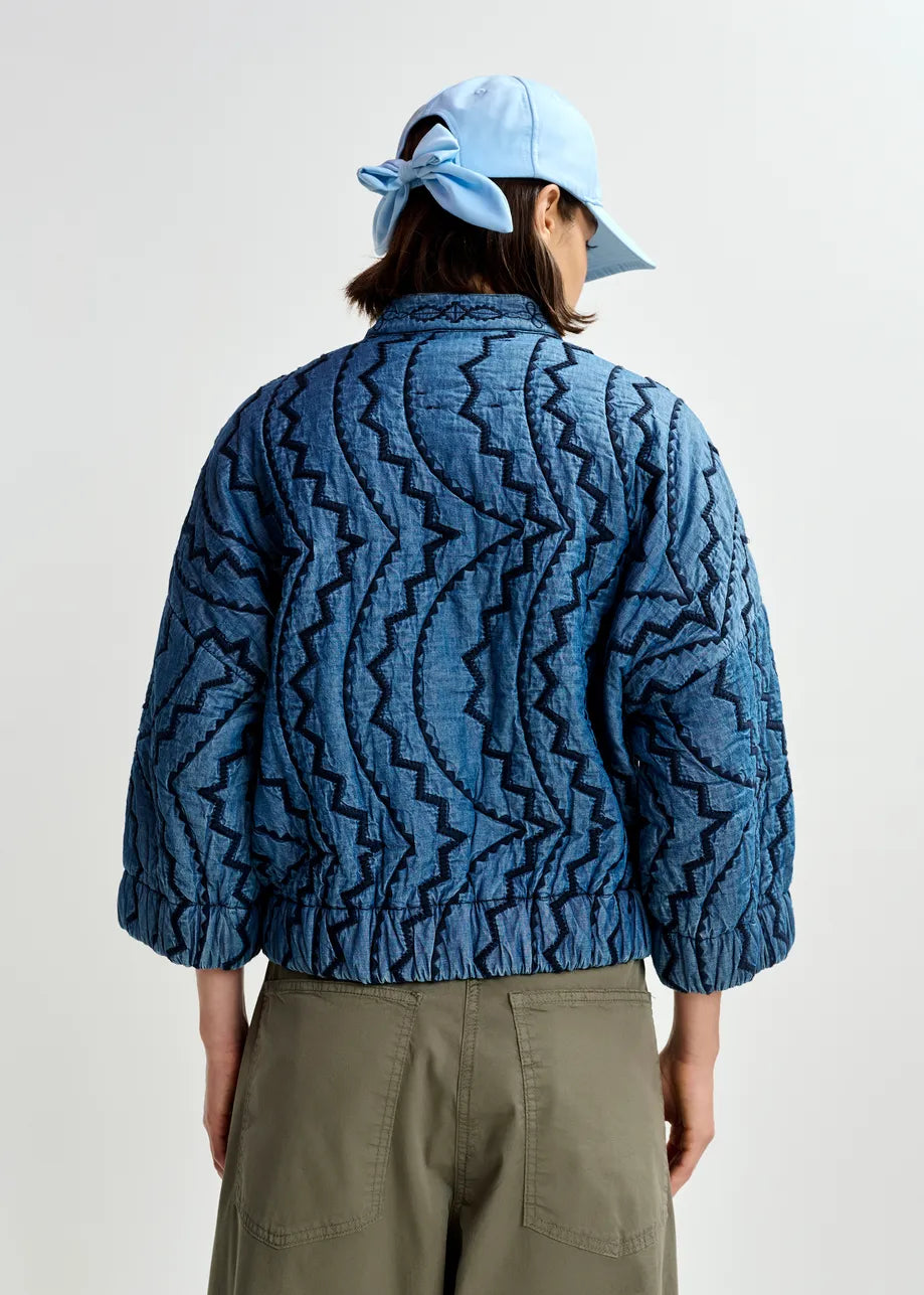 A person faces away, wearing the Essentiel Antwerp Quilted Denim Jacket in blue, paired with olive-green pants and a light blue cap with a bow at the back, set against a plain background.