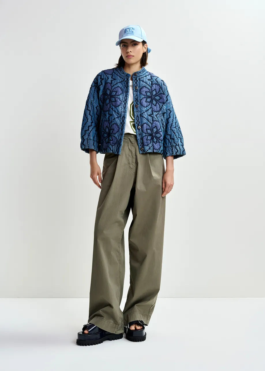 A person wears an Essentiel Antwerp Quilted Denim Jacket in blue, paired with a light blue cap, wide-leg khaki pants, black platform sandals, and a white printed shirt against a plain light background.