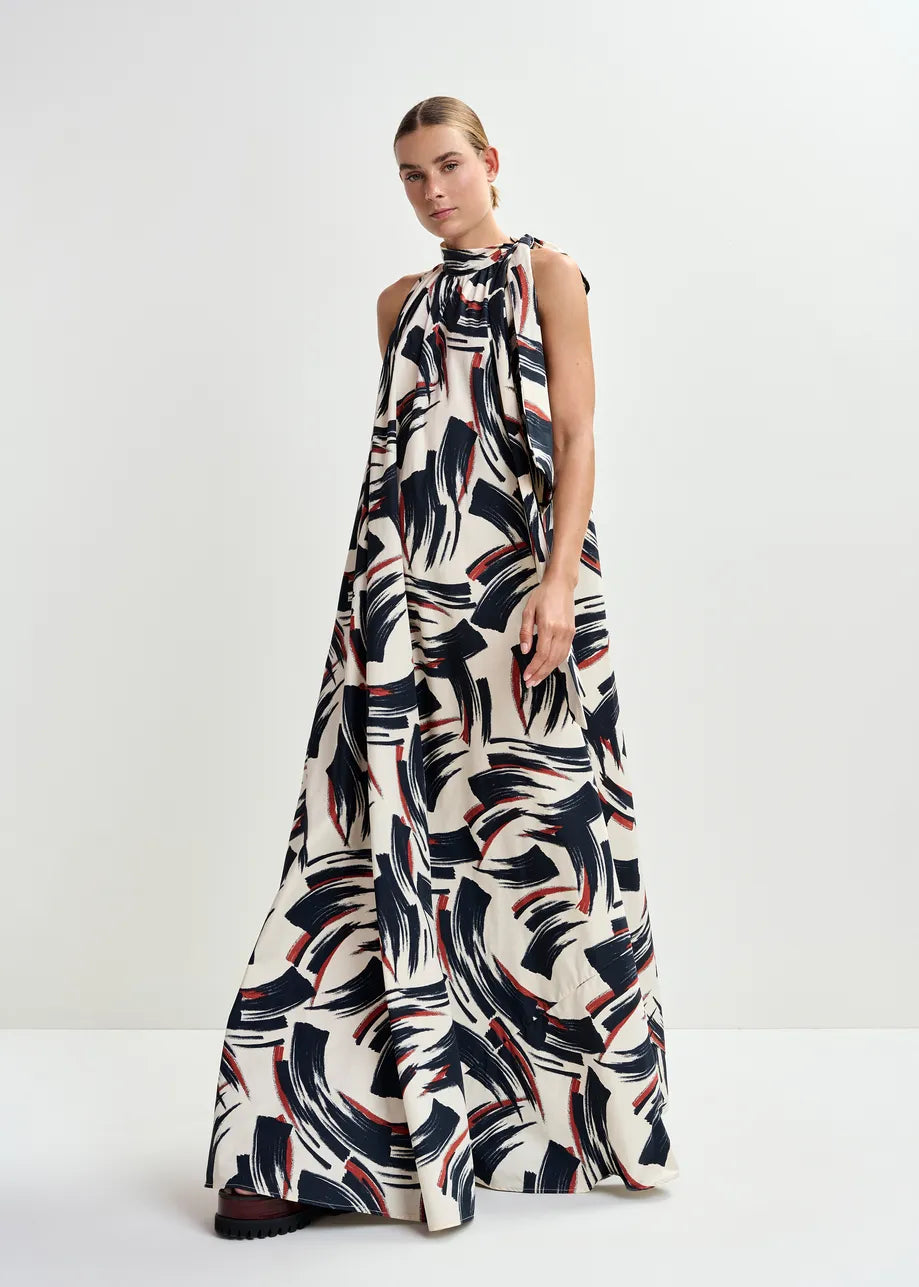 A person stands against a plain white background wearing the Essentiel Antwerp Jayda High Neck Maxi Dress in Burned Red, featuring a recycled-polyester maxi silhouette with a bold black, navy, and red brushstroke pattern and knotted neckline.