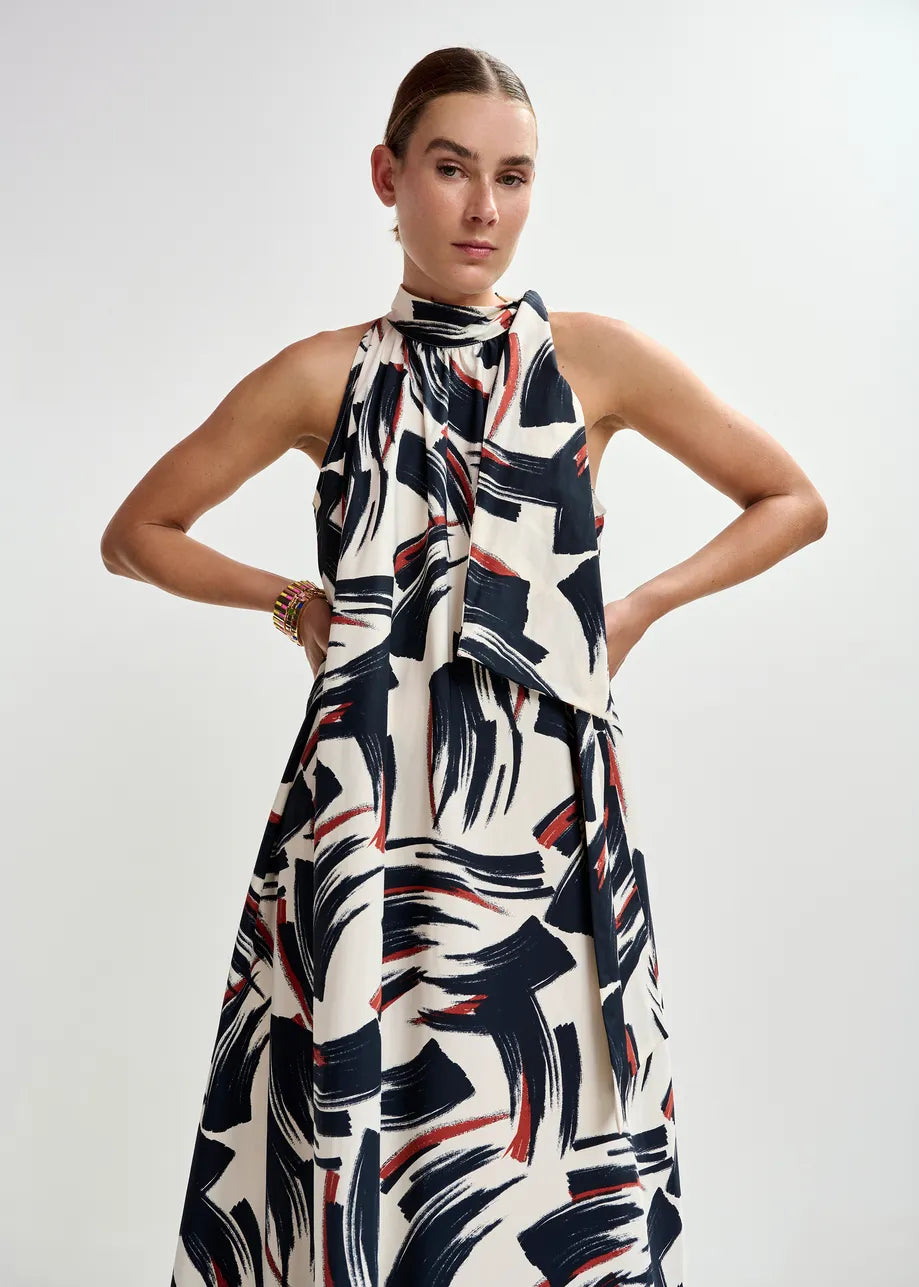 A person stands with hands on hips, wearing the Essentiel Antwerp Jayda High Neck Maxi Dress in Burned Red, featuring a sleeveless design, knotted high neckline, and bold abstract print against a plain light background.