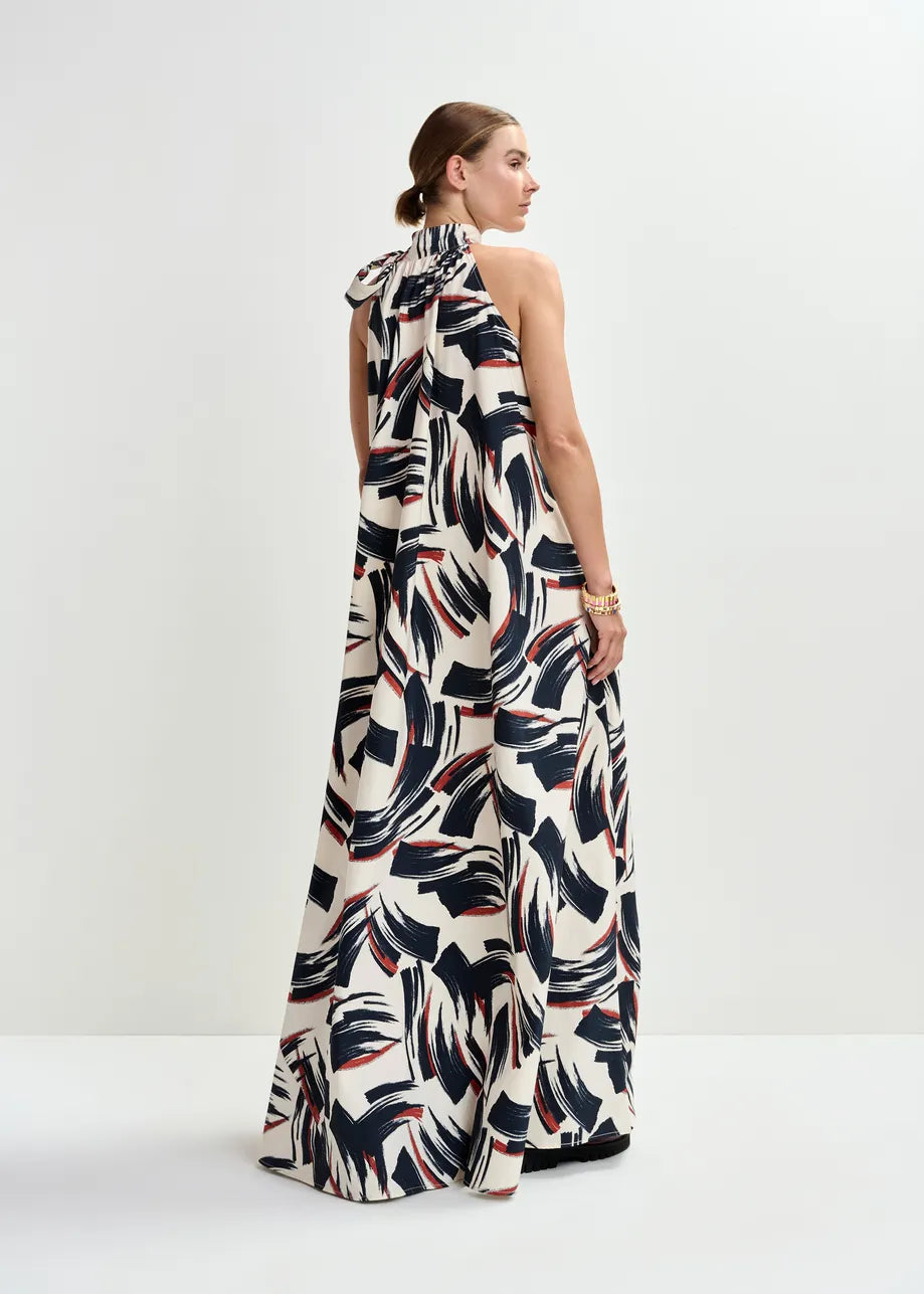 A woman stands in a studio, seen from the back, wearing the Essentiel Antwerp Jayda High Neck Maxi Dress in Burned Red—a sleeveless recycled-polyester maxi with a bold black, navy, and red abstract print and an A-line silhouette.