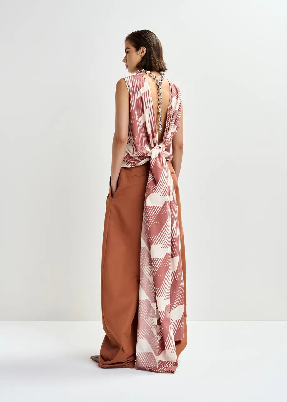 A woman stands against a plain background wearing Essentiel Antwerp’s Jelani Sleeveless Bow Top in Etruscan Red, featuring a cotton wrap design, long draped back tie, and chain detail along the spine, paired with wide-legged brown pants.