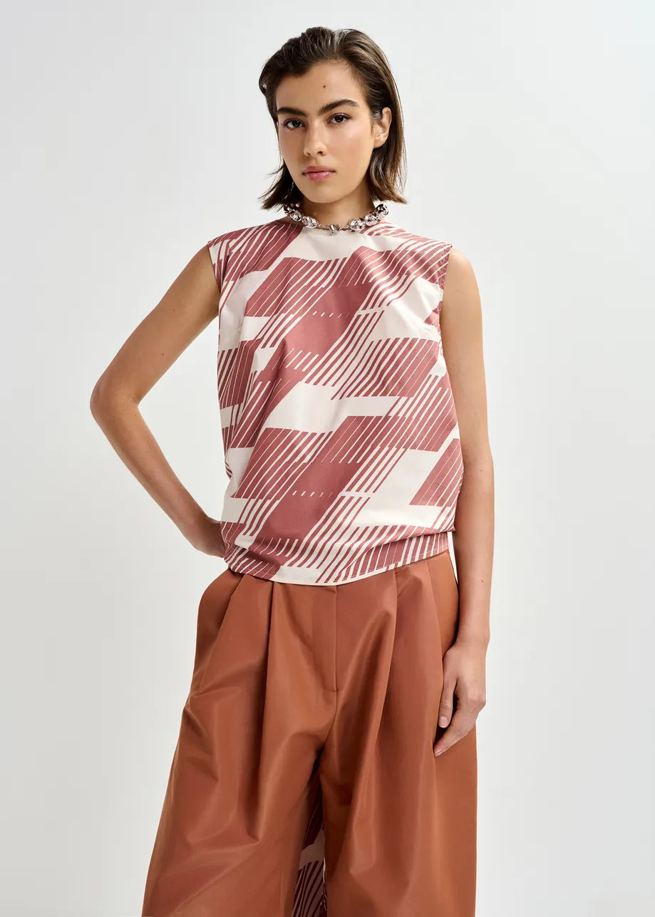 A person wears the Essentiel Antwerp Jelani Sleeveless Bow Top in Etruscan Red, paired with wide-leg, high-waisted rust-colored pants, standing against a plain light background.