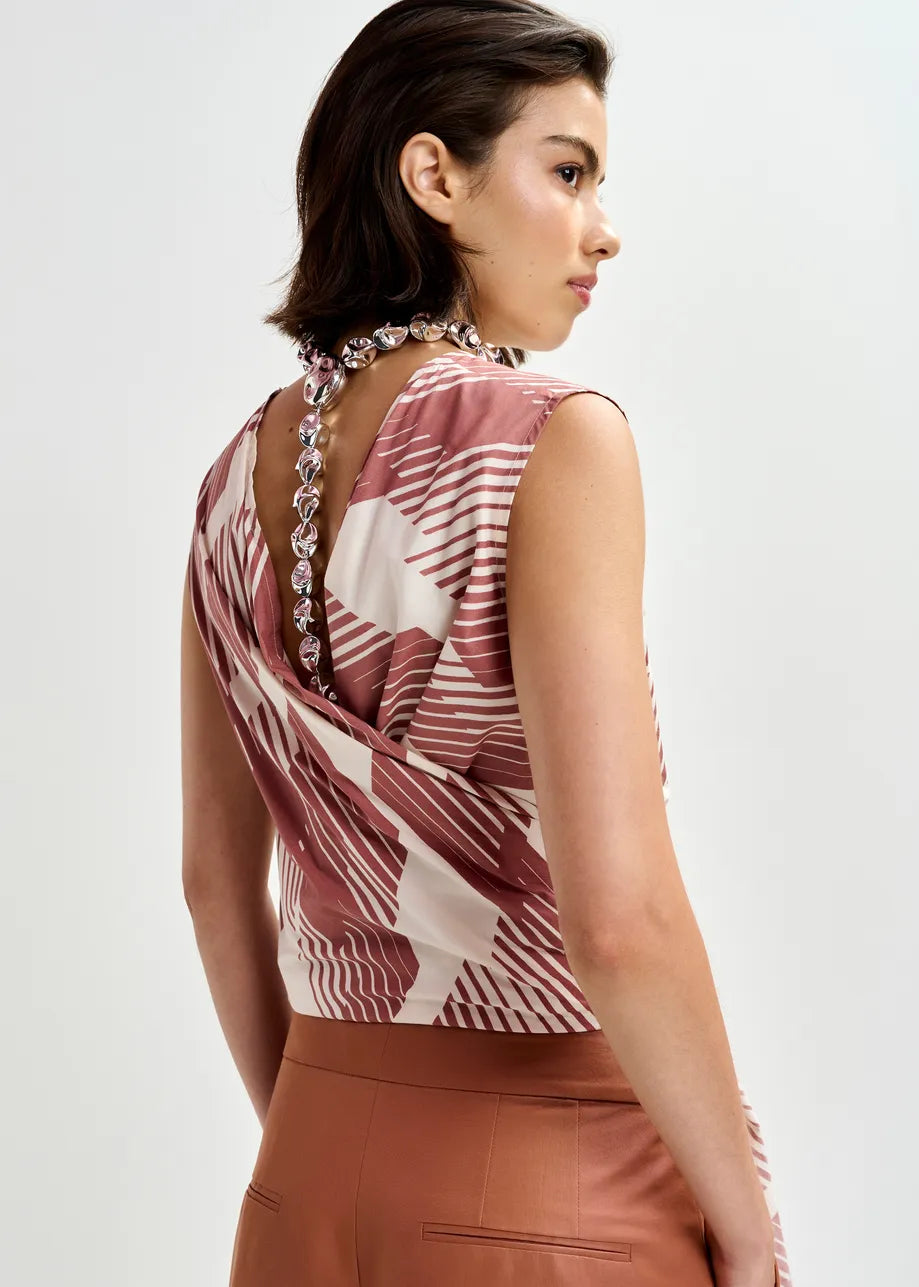 A woman with short brown hair and a beaded necklace wears the Essentiel Antwerp Jelani Sleeveless Bow Top in Etruscan Red, featuring a wrap design and draped fabric, paired with brown pants.