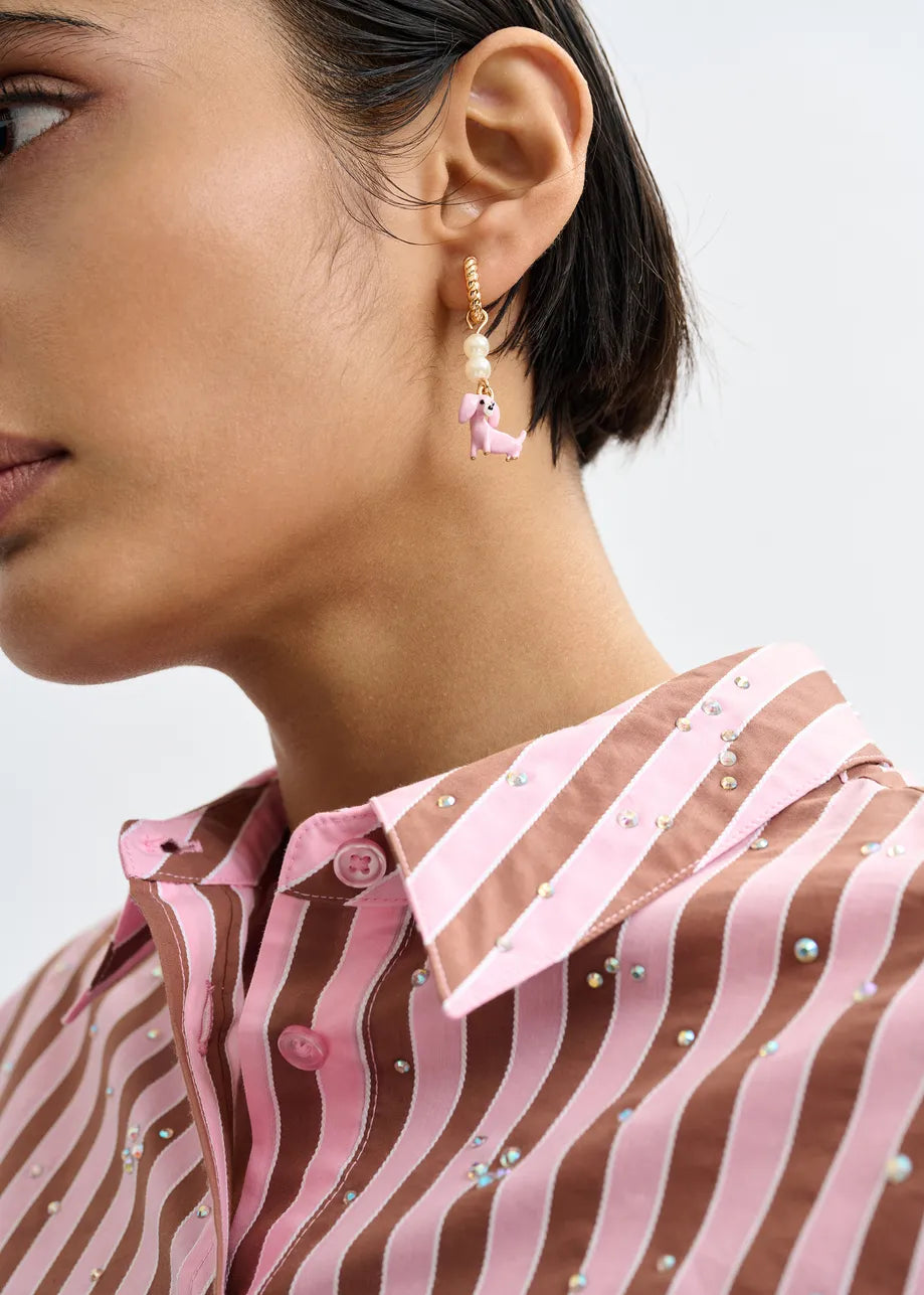 Close-up of a person in a pink and brown striped shirt with rhinestones, wearing Essentiel Antwerp's Gold Drop Earrings - Pink Dachshund and another unique style, with short dark hair partially visible.