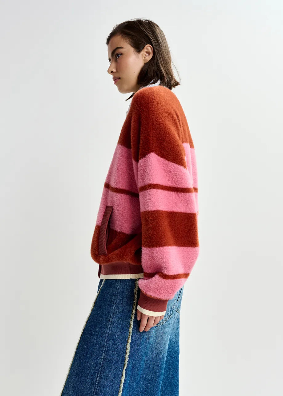 A person wearing the Essentiel Antwerp Jicama Knitted Bomber in red and pink with blue jeans stands against a plain light background.