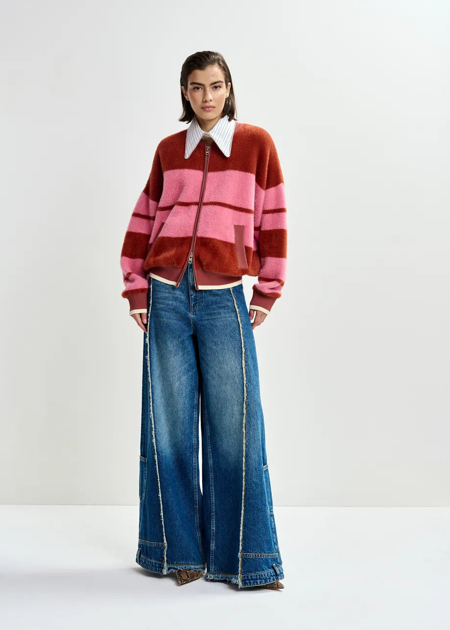 A woman stands against a plain background, wearing the Essentiel Antwerp Jicama Knitted Bomber in red and pink over a collared shirt, paired with wide-leg blue jeans featuring frayed seams.