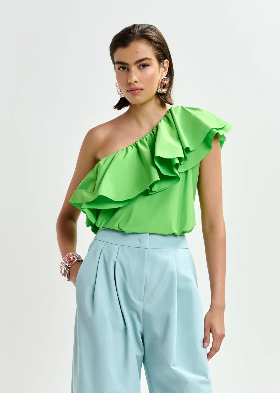 A woman wears the Essentiel Antwerp Jillian Asymmetric Ruffle Top in green with light blue high-waisted trousers, standing against a plain white background.