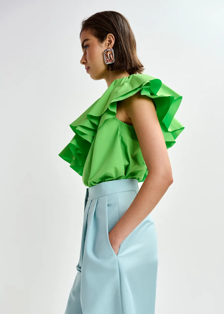 A woman in profile wears the Essentiel Antwerp Jillian Asymmetric Ruffle Top in bright green recycled polyester, paired with light blue high-waisted pants. Her short brown hair and geometric earrings pop against a plain light background.